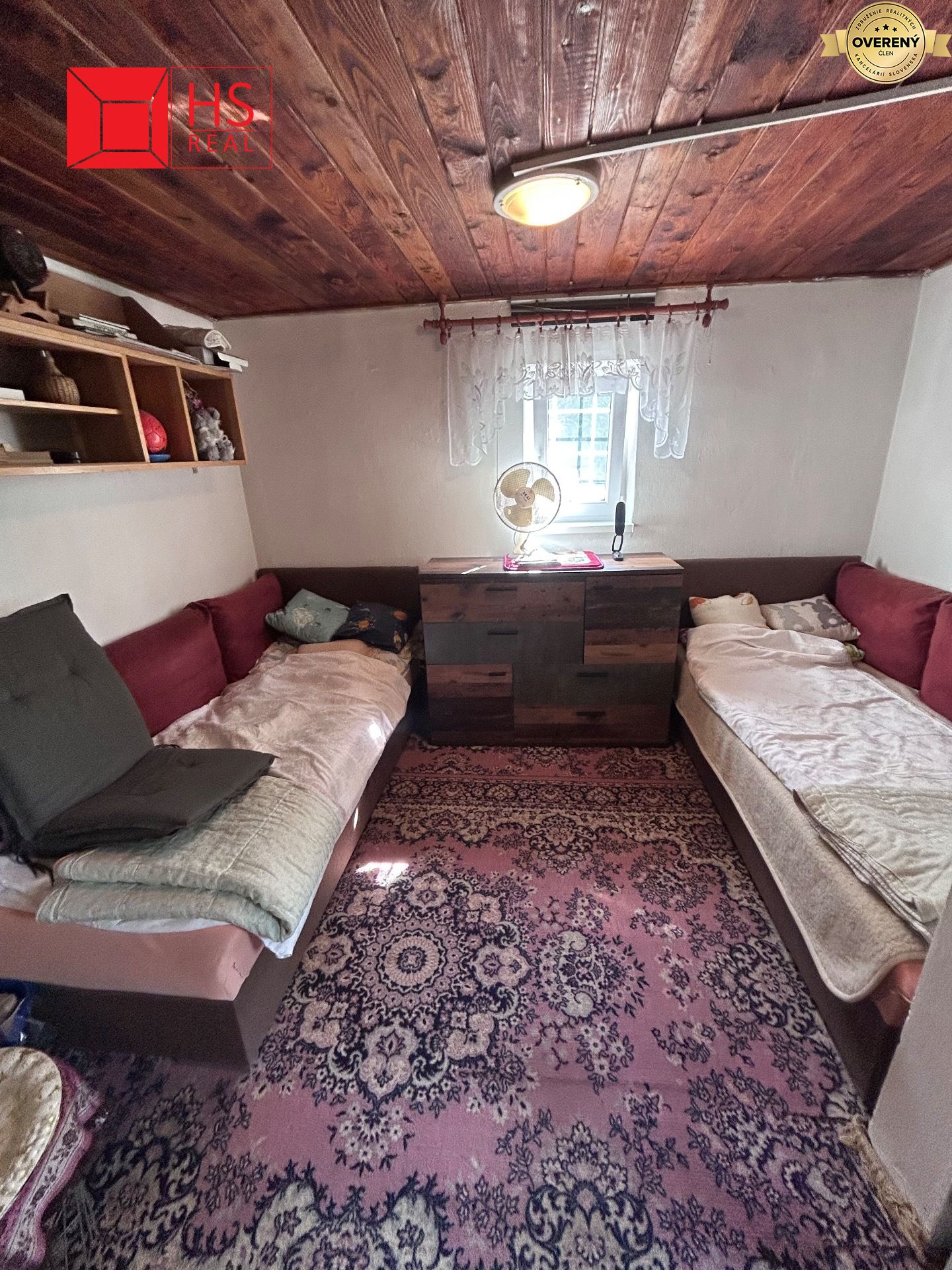 Chata s dvomi lôžkami, drevený obklad stropu, koberec a komoda pri okne.